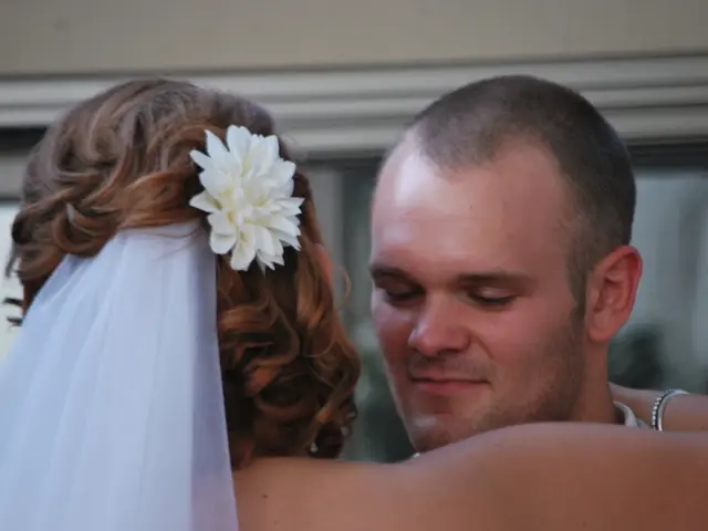 Here I can see a woman hugging a man. On her head, I can see a white color flower and a scarf. In...